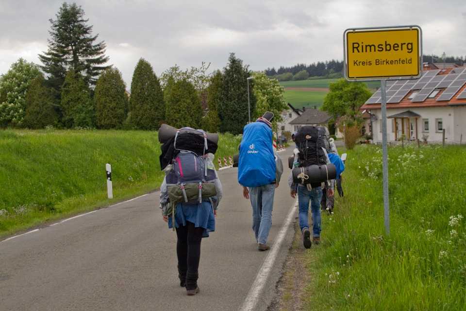 Pfadfinder laufen mit Rucksäcken an einem Ortsschild vorbei